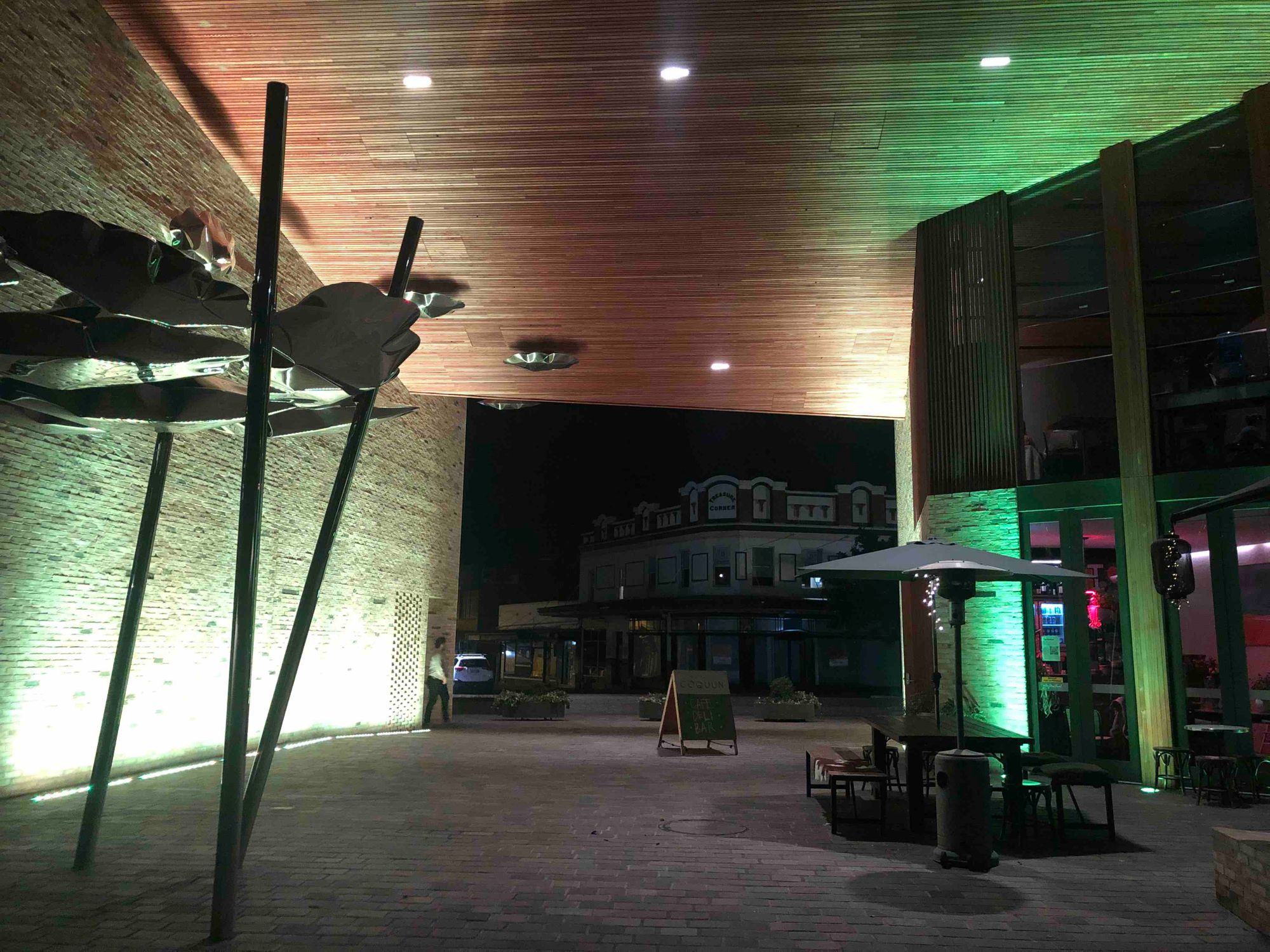 A night tunnel, with a high wooden roof lit up in green, with stone walls either side, and restaurant tables in the middle.