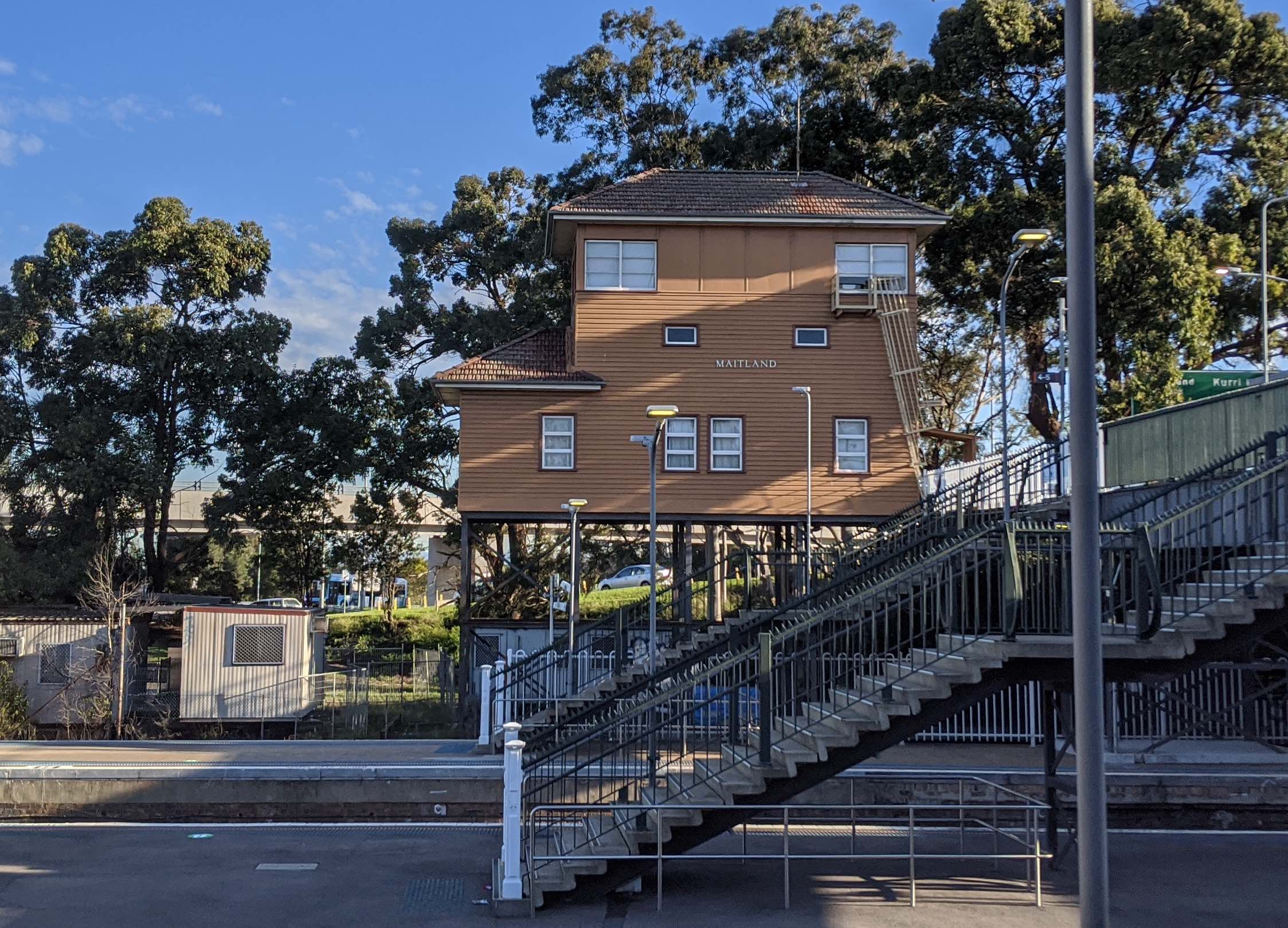 Background: a two-story wooden house, about 8m high up on stilts off the ground, marked "Maitland". Train platform foreground.