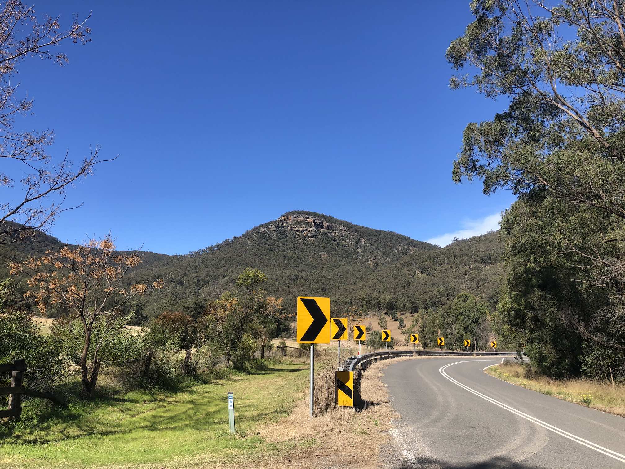 Windy road on right, farm to the left, sandstone mountain set against blue sky.